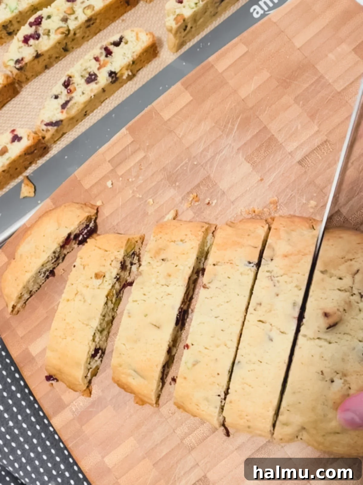 Vibrant Cranberry Pistachio Biscotti 5 Hands slicing a baked biscotti log on a wooden cutting board with a serrated knife.