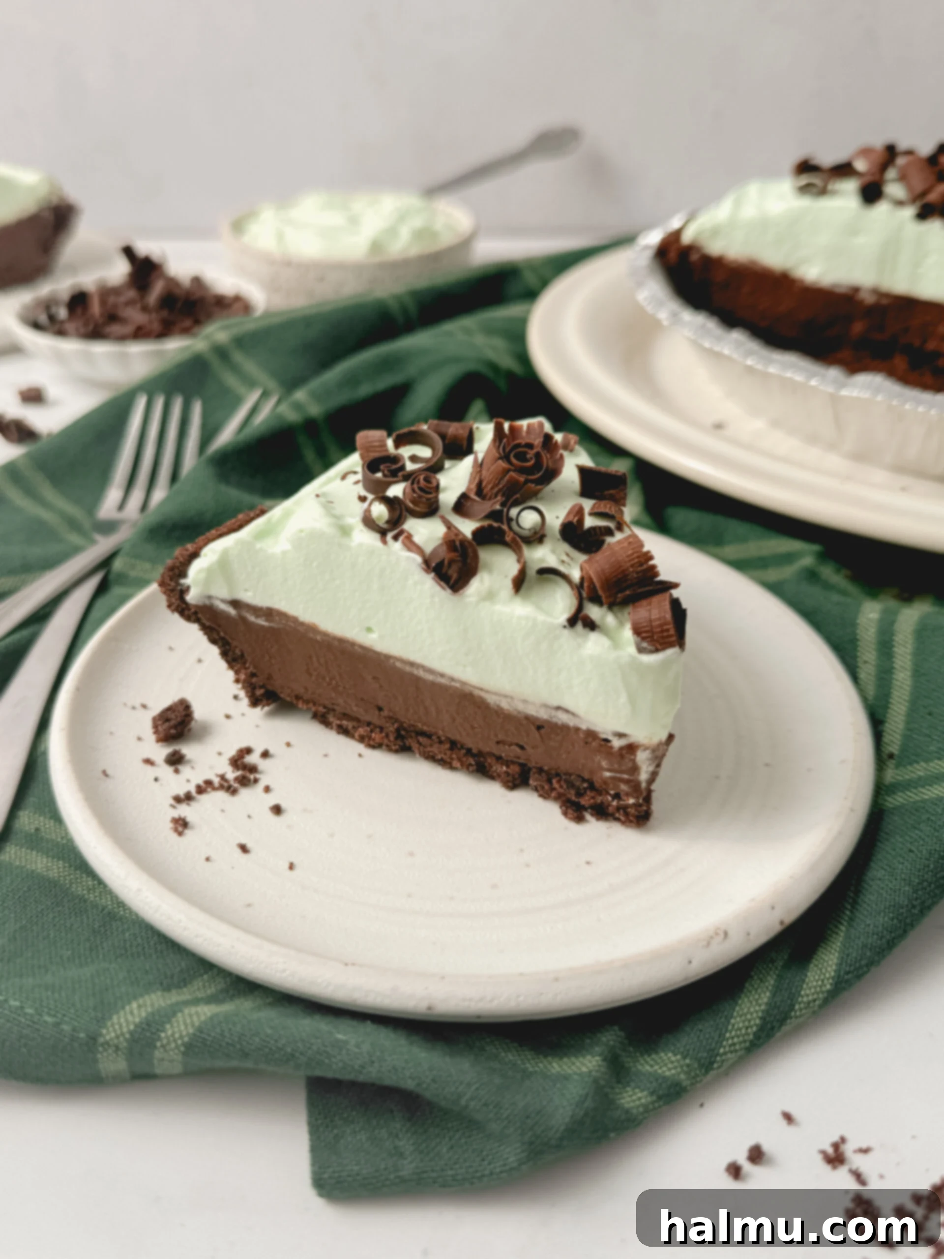 A slice of Chocolate Mint Cream Pie on a white plate, showcasing its layers of chocolate cookie crust, creamy chocolate custard, and luscious minty whipped cream, topped with chocolate curls.