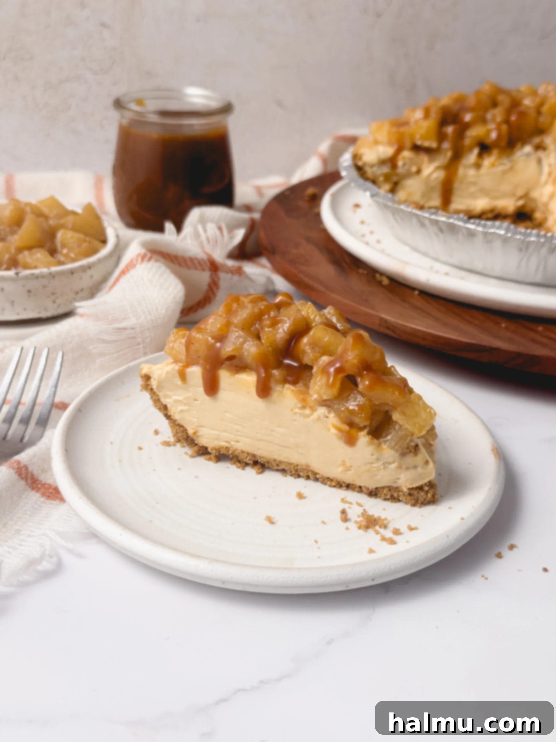 A slice of Caramel Apple Cheesecake Pie on a plate, showing layers of graham cracker crust, cheesecake, and apple compote, drizzled with caramel sauce.